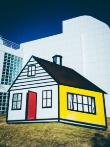 Cartoon-style house sculpture against modern building facade on a sunny day.